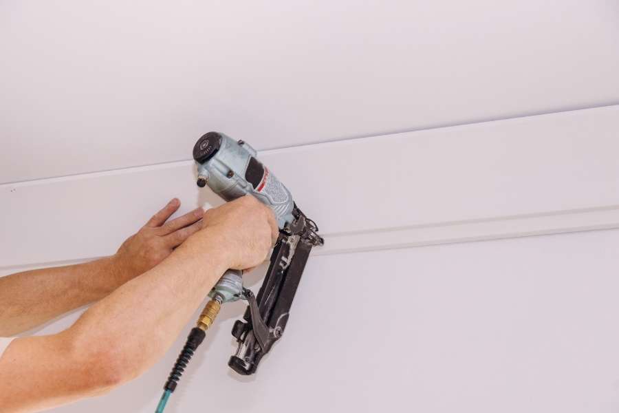 A worker using a pneumatic nail gun to secure white crown molding along the junction of a wall and ceiling.
