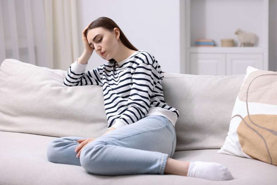 A young woman sitting on a light-colored sofa holding her head, experiencing digital fatigue from excessive screen time.