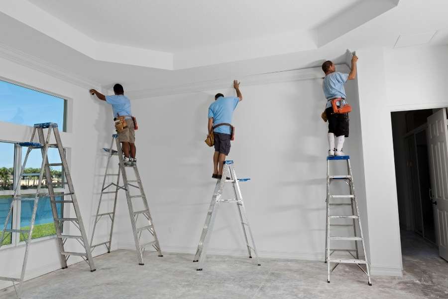 Three professional contractors standing on separate ladders installing white crown molding in a new room.