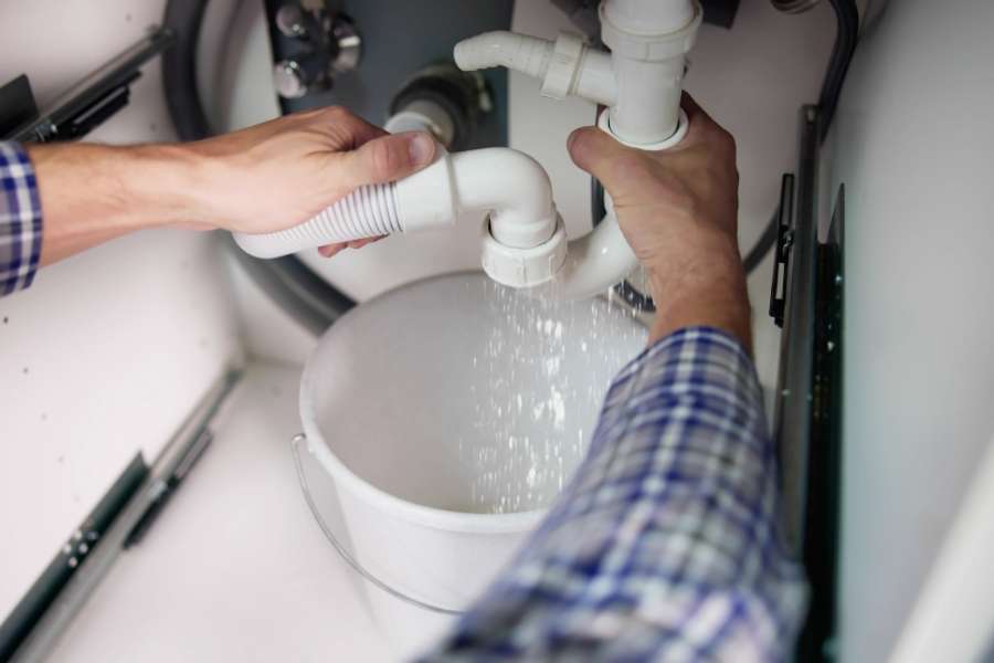A person in a plaid shirt disconnecting white PVC pipes over a bucket to catch a hidden water leak.